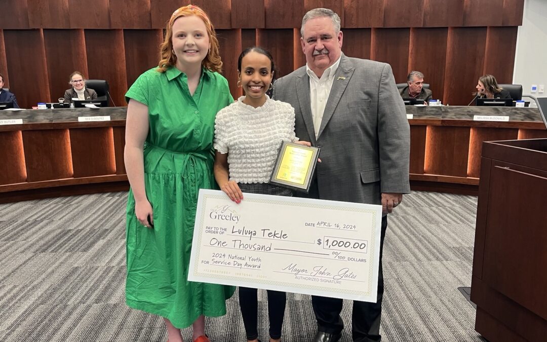 Mayor presenting oversized check in City Council chambers to a female high school student that won the 2024 National Youth Service Day Awards $1,000 scholarship from the Greeley Youth Commission Chair.